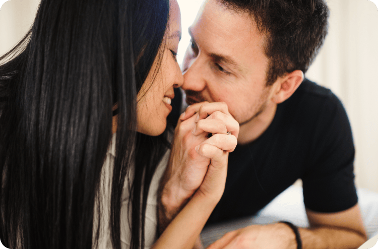 Couple close together in bed, woman smiling as man kisses her hand gently