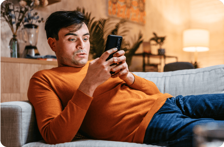 A man lying on a couch, looking at his smartphone with a serious expression.