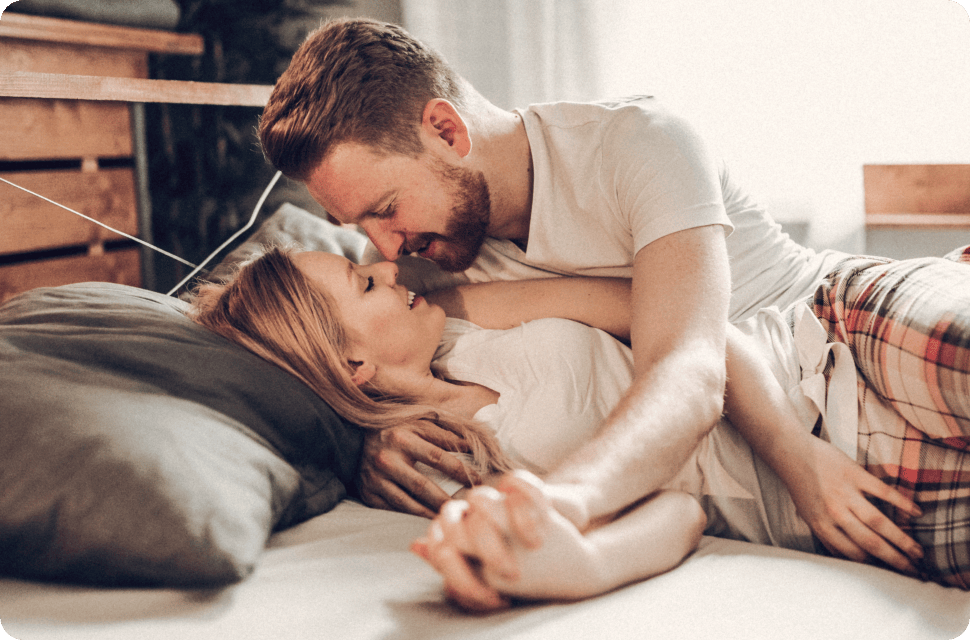 A couple lying in bed, embracing and gazing at each other tenderly.