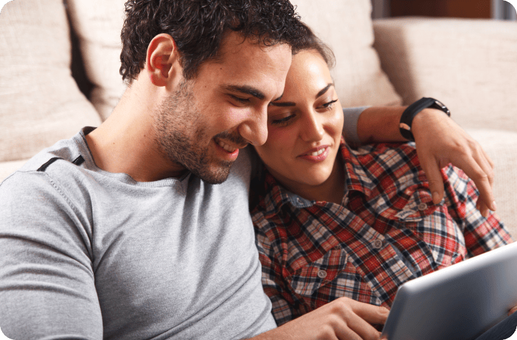 A couple sitting on a couch, leaning on each other and looking at a tablet together.