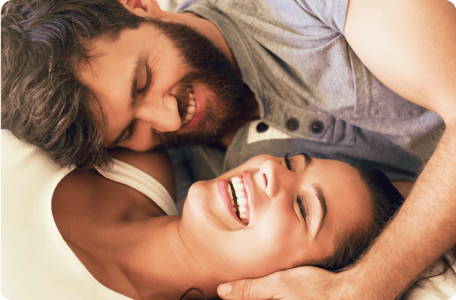 Bearded man in a gray shirt, smiling widely as he lies on top of a laughing woman in bed