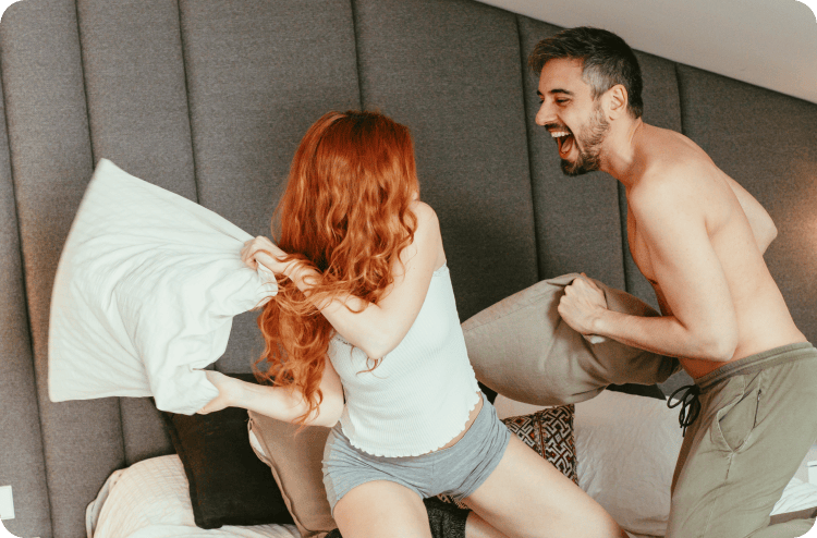 A joyful pillow fight between a man and a woman on a bed, both laughing in a playful and lighthearted moment