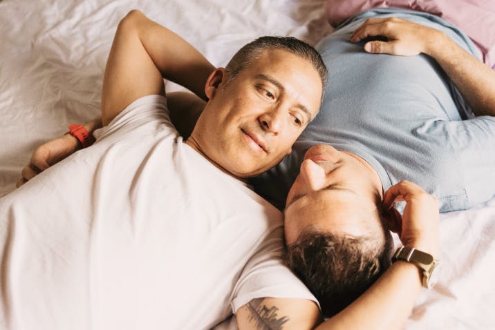 Two men lying in bed, smiling and looking at each other affectionately