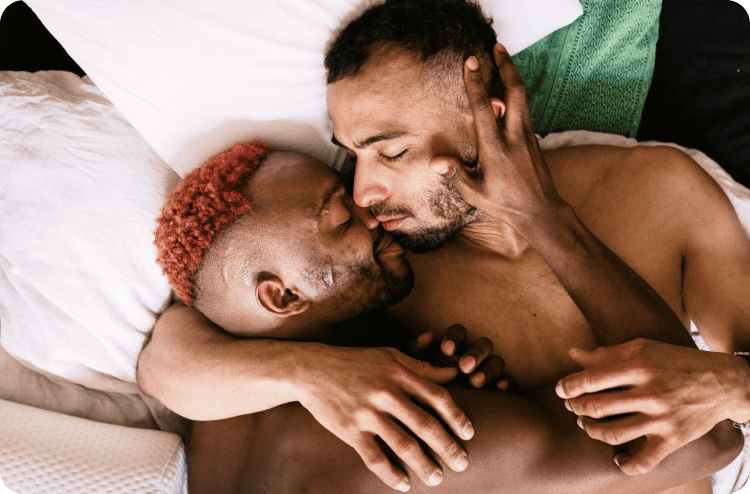 Two men in bed, one kissing the other's cheek tenderly, both shirtless and intimate