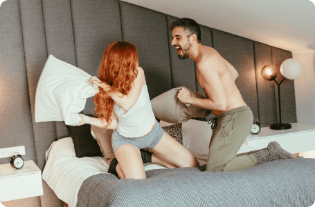 A couple having a playful pillow fight on a bed, both laughing and enjoying the moment.